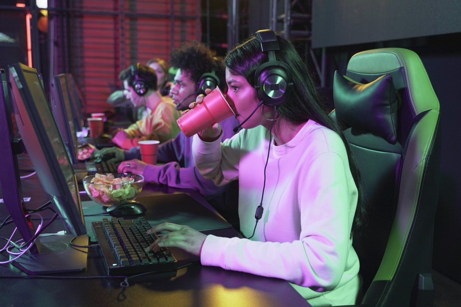 Players at a row of gaming PCs in a neon-lit esports café, with drinks and snacks on the desk.